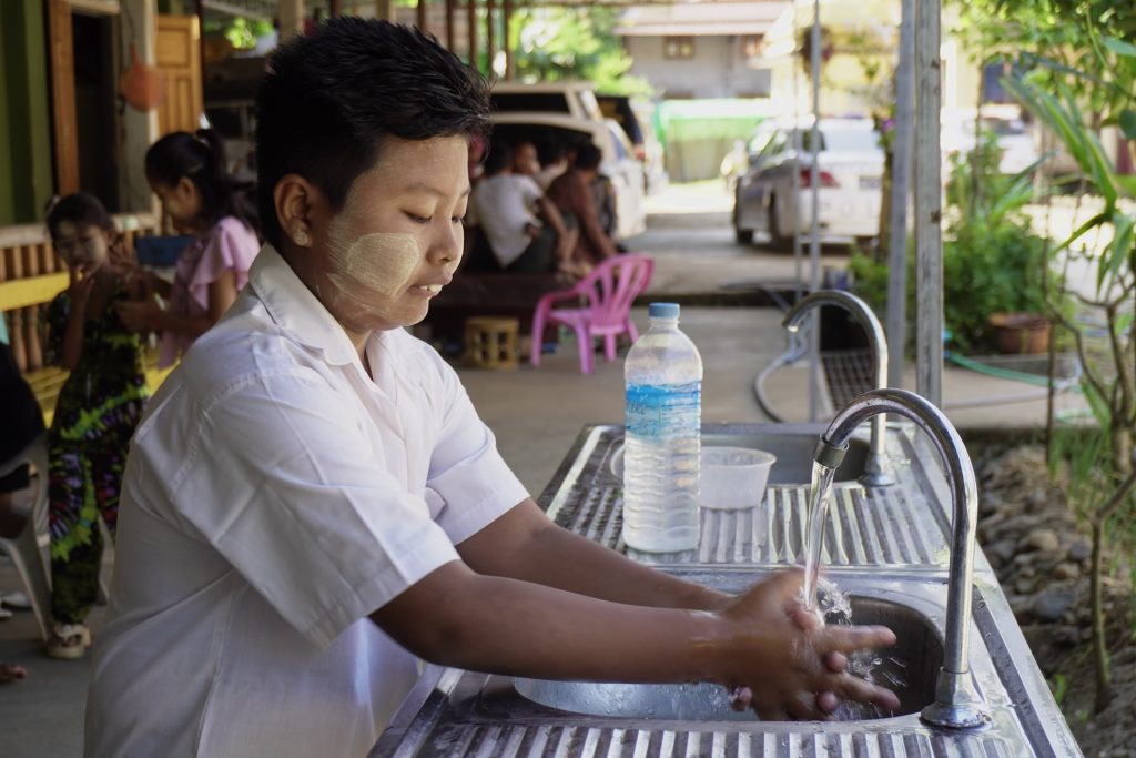 A boy with Thanaka on his face washes his hands at an outside basin.