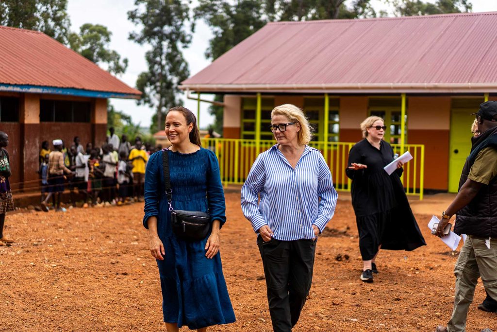 Kaksi suomalaista kansanedustajaa kävelee Ugandassa pakolaisalueen koulun pihalla. Taustalla on muita ihmisiä ja koulun rakennuksia.
