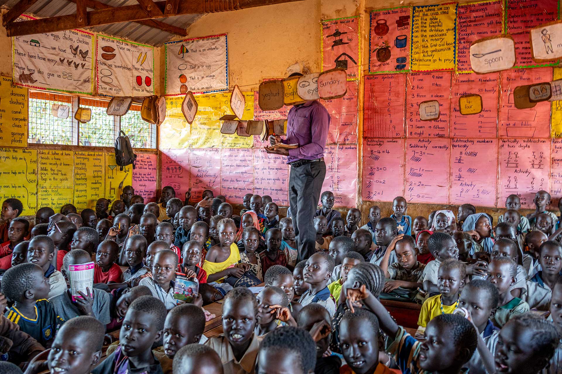 Opettaja kävelee pakolaisalueen lapsia täynnä olevassa luokkahuoneessa pulpettien päällä.