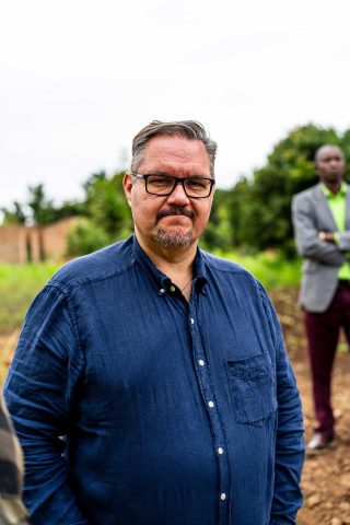 Ulkona seisova mies katsoo kameraan. Taustalla näkyy toinen mies.