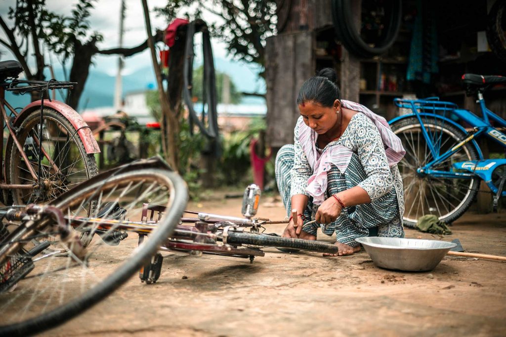 Nainen istuu kyykyssä maassa korjaamassa polkupyörää Nepalissa. Taustalla näkyy muitakin polkupyöriä. 