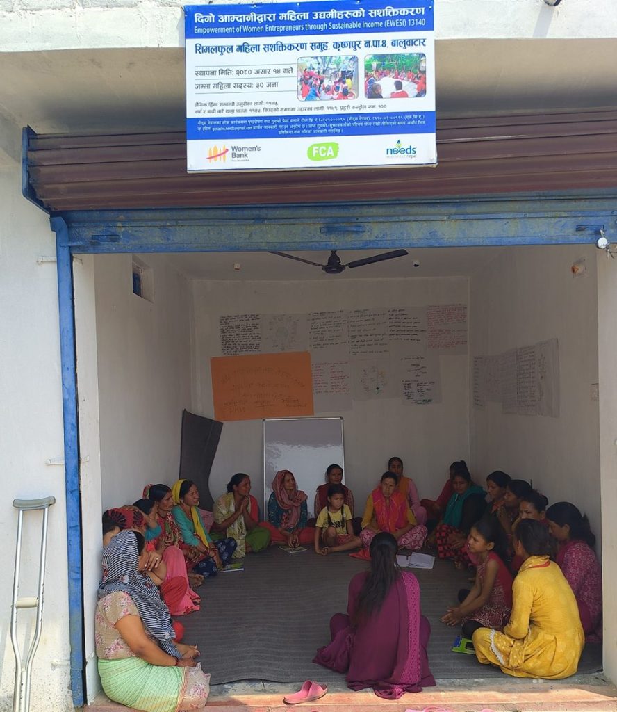 A group of women sit in a circle in a small room. A sign above the doorway shows the logos of Women's Bank, FCA and Needs (a local Nepali organisation)