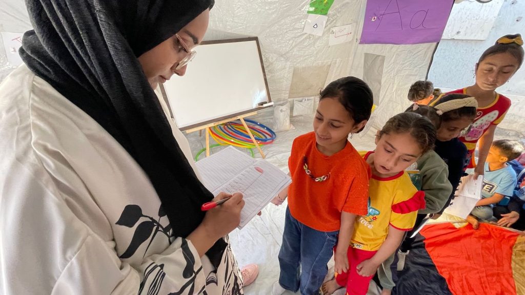A teacher writes in a book while children line up in front of her. The setting is recognisable as a classroom with whiteboard and playthings, but the walls are canvas.