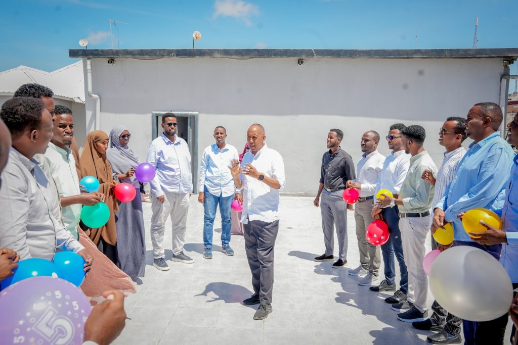 A number of people stand outside in a circle. Some are holding balloons. They are listening to a man speaking in the middle of the circle.