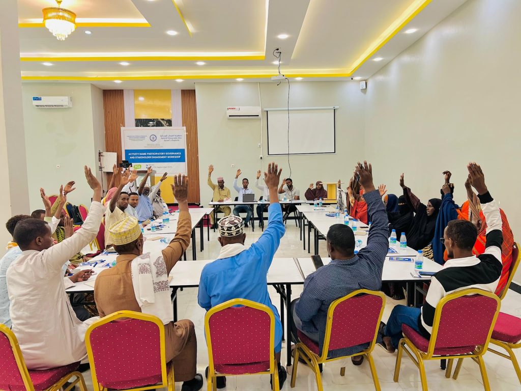 People with their hands raised sit around tables arranged in a circle in a conference room.