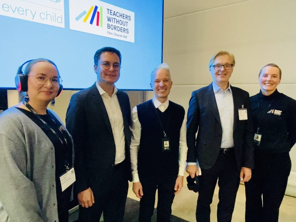 A group of people smile for a photograph while standing in front of a screen displaying the logos of UNICEF and Teachers Without Borders