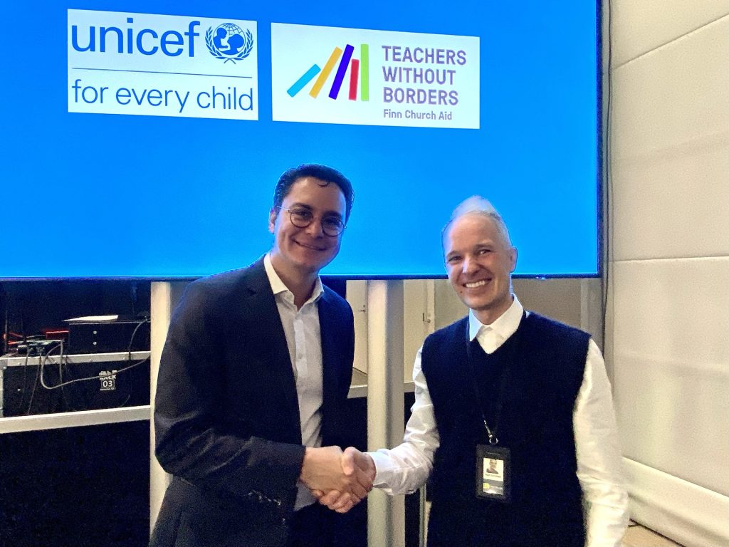 Two men shake hands in front of a screen displaying the logos of UNICEF and Teachers Without Borders