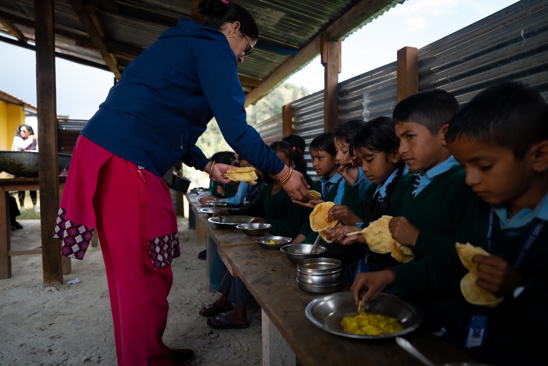 A line of young primary school children in uniforms sit at a long bench and receive naan from their teacher.