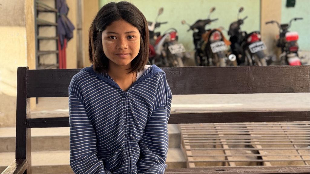 A smiling teenager sits on a school bench and looks into the camera.