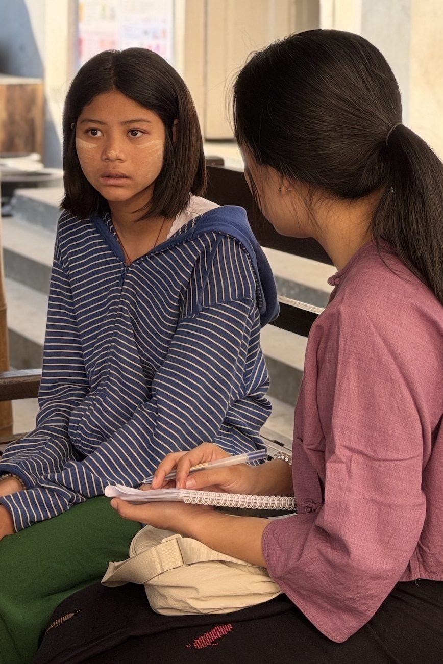 A teenaged girl sits on a bench. An older woman sits next to her and speaks.