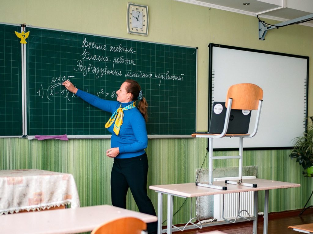 Opettaja kirjoittaa liitutaululle luokkahuoneessa Ukrainassa. Opettajan edessä on tuoli, jonka päälle nostetun kannettavan tietokoneen kautta opettaja pitää oppitunteja lapsille, jotka eivät voi sodan takia tulla kouluun. 