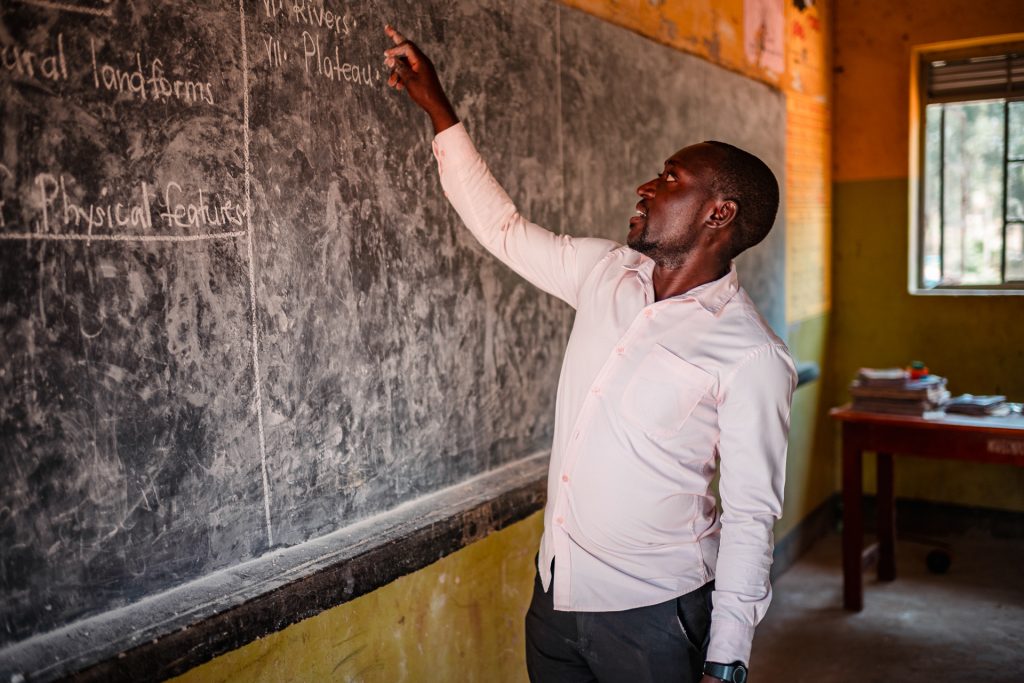 A teacher stands at a blackboard and points to a written sentence