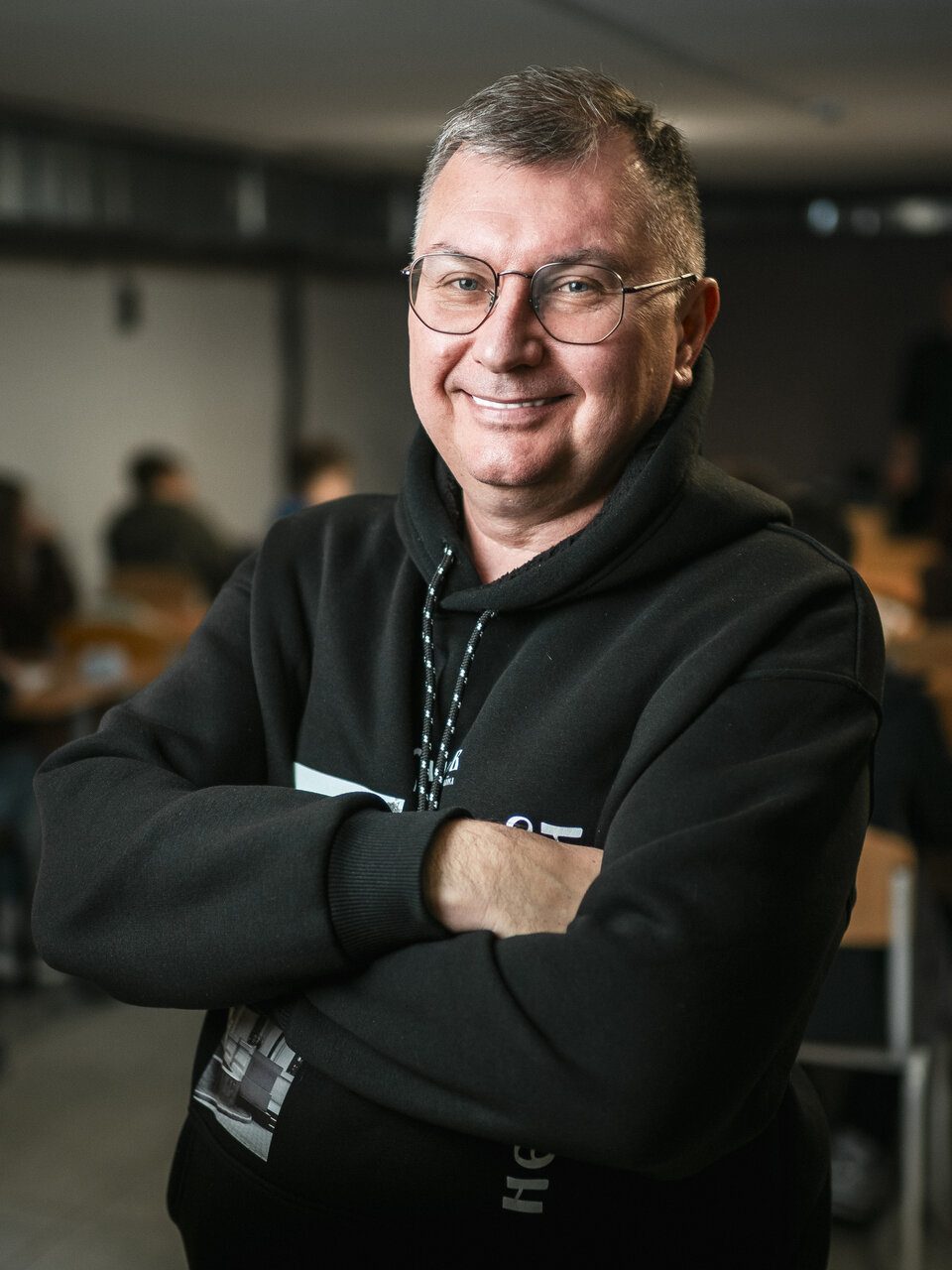 A smiling man in glasses and wearing a black hoodie looks directly into the camera. Behind him in the background is a classroom.