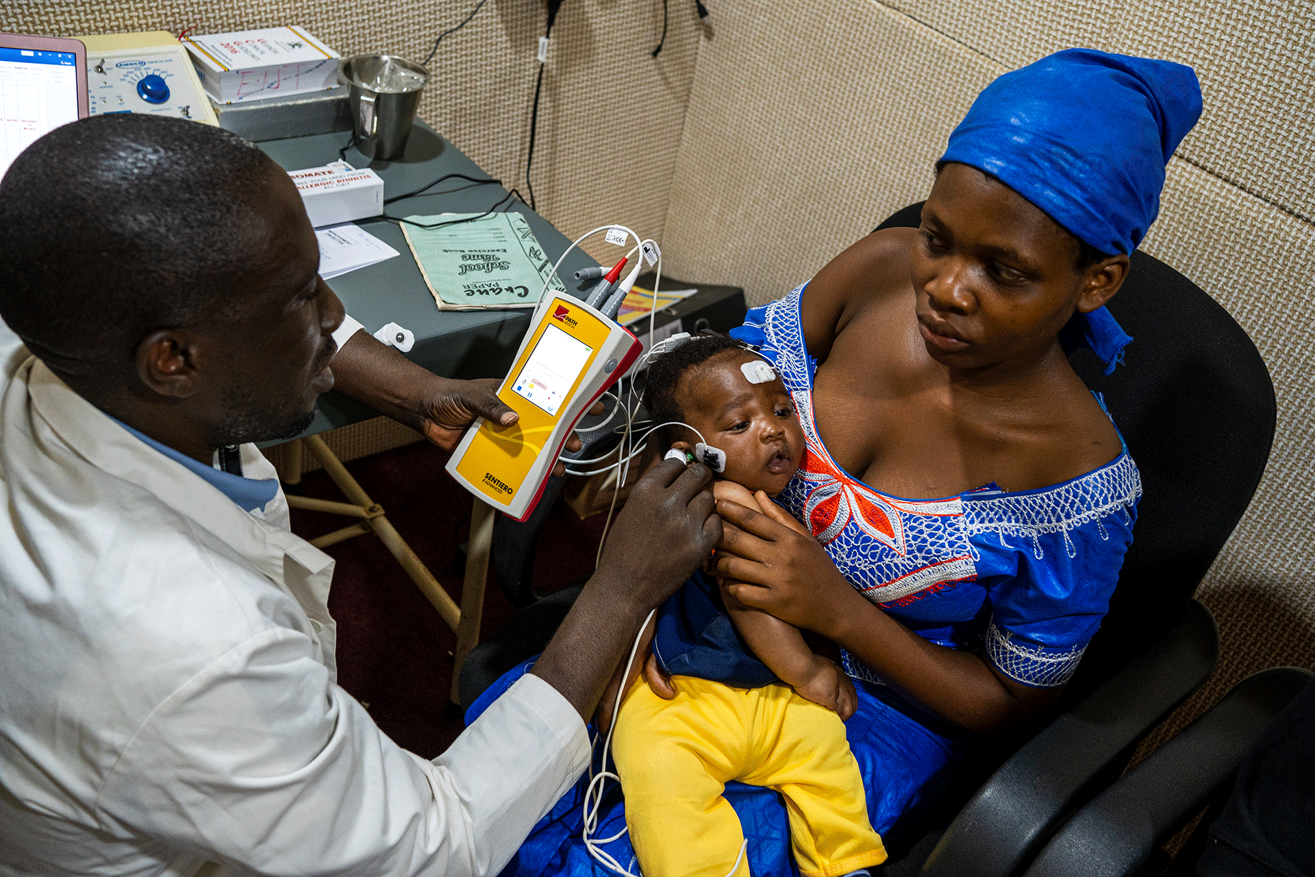 Lapsi makaa äitinsä sylissä kun lääkäri suorittaa hänelle kuulotestiä.