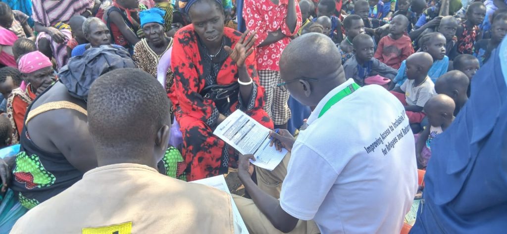 A man with a clipboard is taking details while a woman in speaking to him standing in front of a large crowd of seated people.