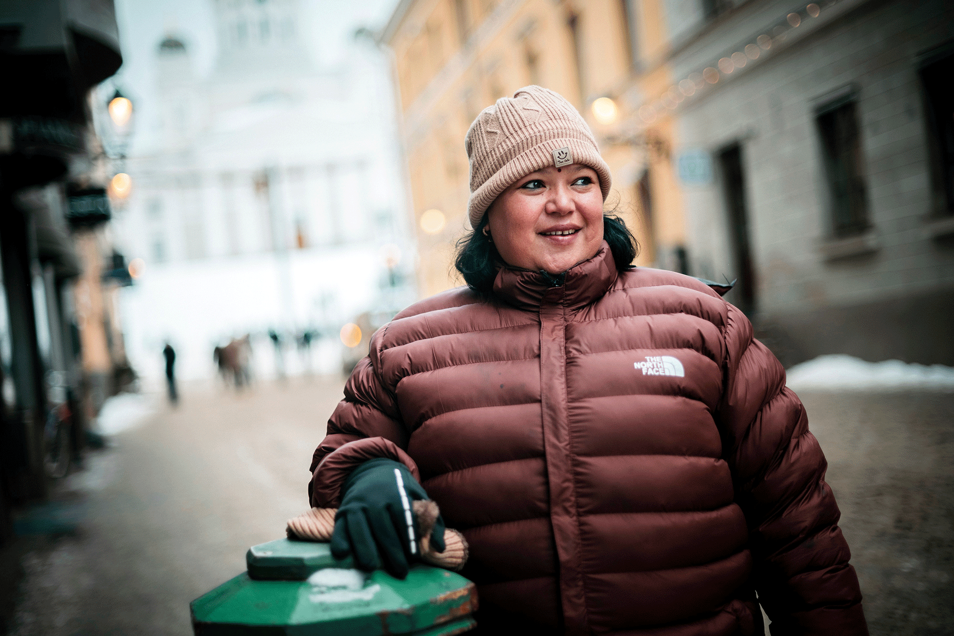 Nainen pipo päässään ja toppatakki päällään nojaa vihreään pylvääseen.