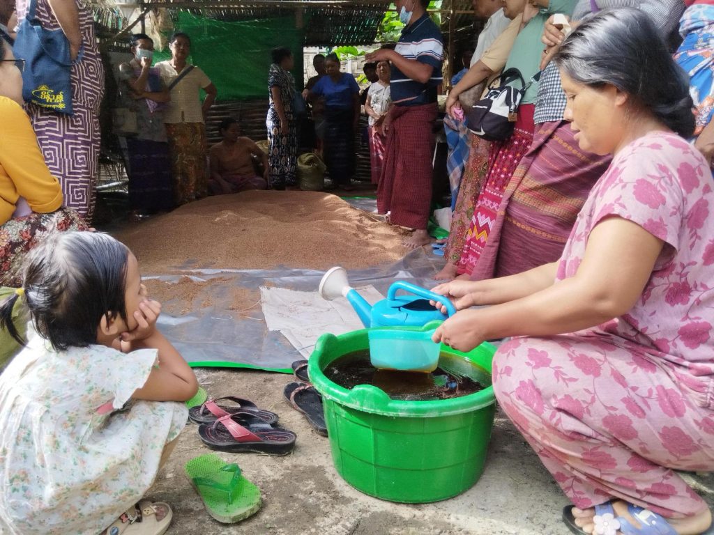 A woman and a small child sit on the ground. The woman holds a small watering can over a bucket. Around them people are standing and watching