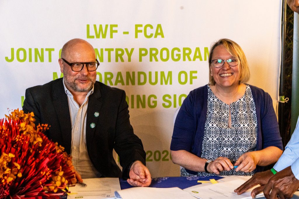 Two smiling people sit at a table. Behind them is a banner with the text, "LWF-FCA Joint Country Programme Memorandum of Understanding Signing"