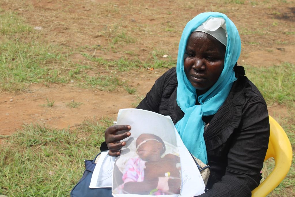 A woman in a blue headscarf sits in a chair and sadly holds her a photo of a child in hospital
