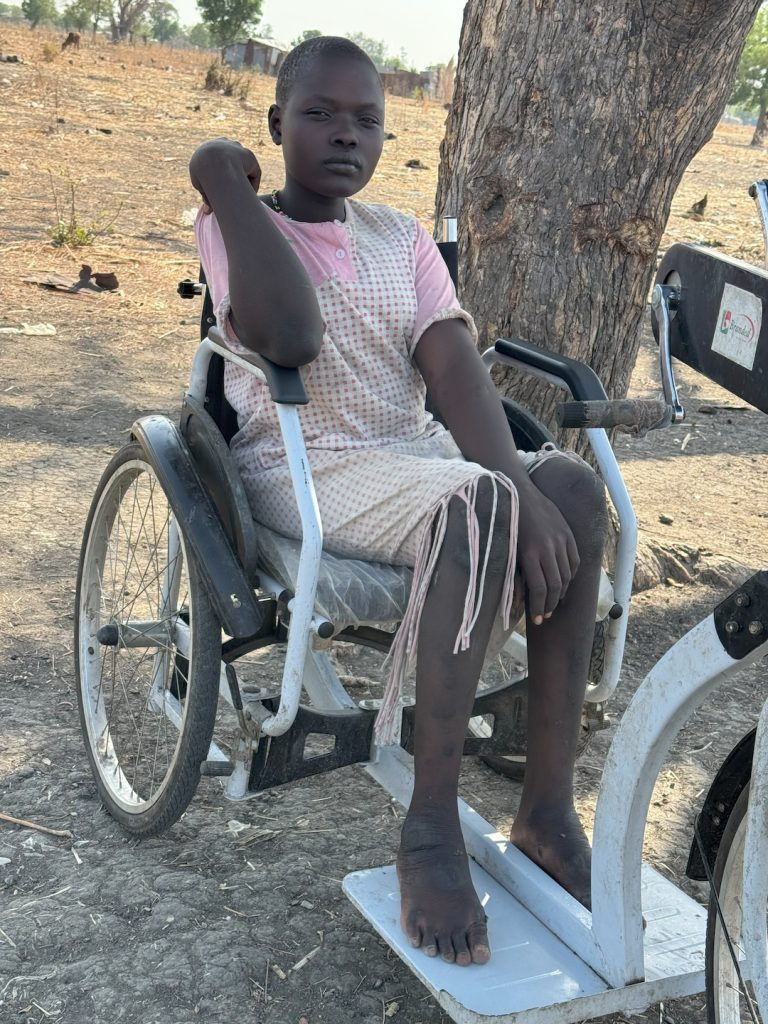 A woman sits in a wheelchair connected to a hand-pedaled bicycle. Her feet are bare.