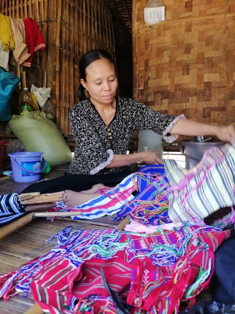 A woman sits on the floor of a bamboo shack and spreads a number of colourful cloths out in front of her.