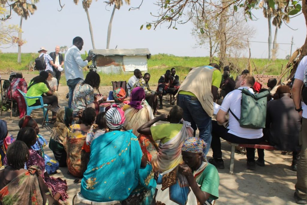 Suuri joukko ihmisiä istuu ulkona varjoisassa paikassa Etelä-Sudanissa. Taustalla näkyy puita ja peltiseinäinen rakennus.