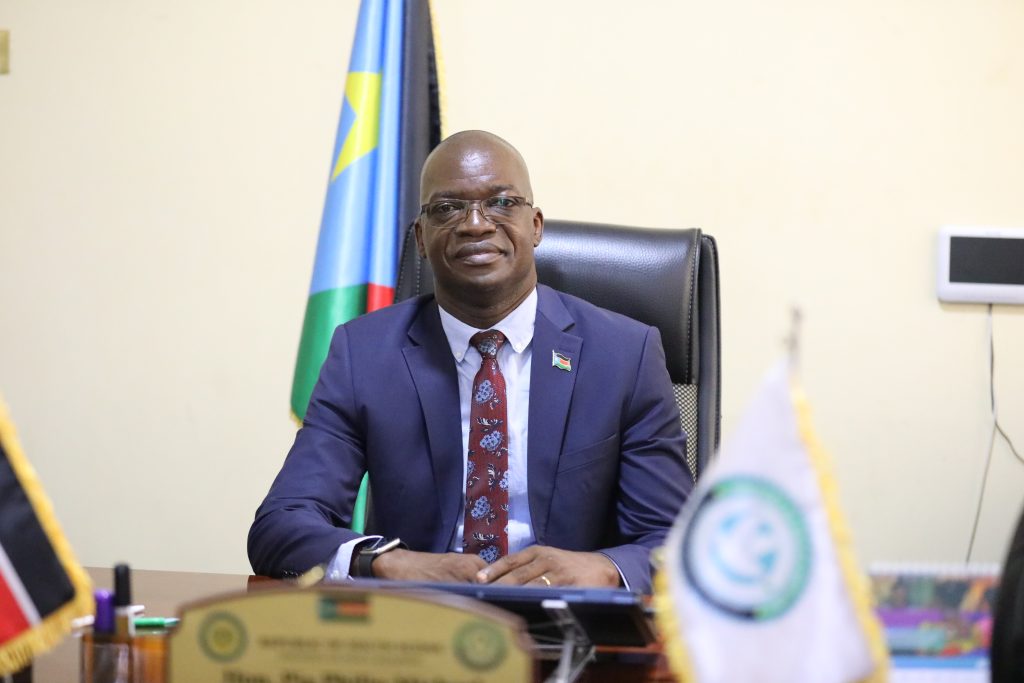 A man in a suit sits at a desk. The flag of South Sudan is behind him. 