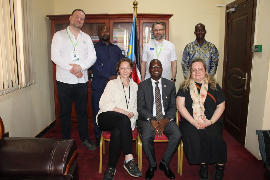 A group of people in an office pose for a photo. The flag of South Sudan is visible in the background.