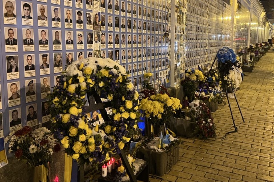 A wall of photos at a memorial in Kyiv. Below the photos there are bouquet and wreaths of flowers.