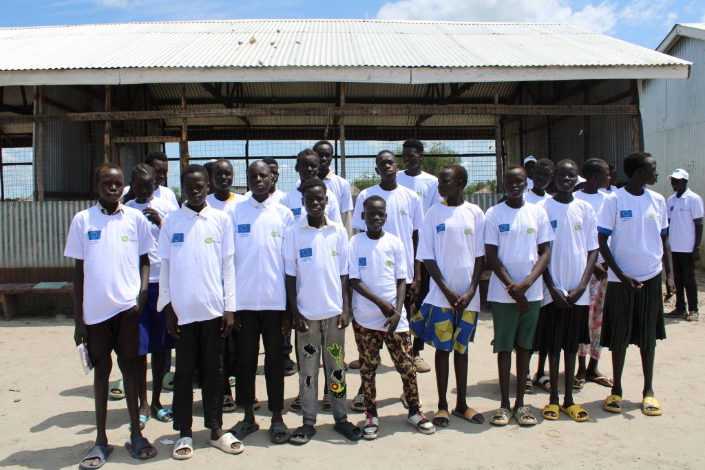 A class-sized group of children pose for a photo outside of a shack. They are wearing white shirts with the EU and FCA's logos.