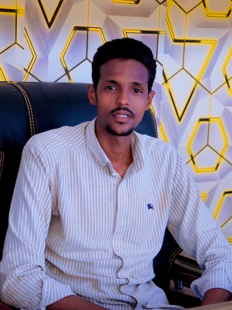 A man in a shirt sits in front of a geometrically patterned wall and looks into the camera.