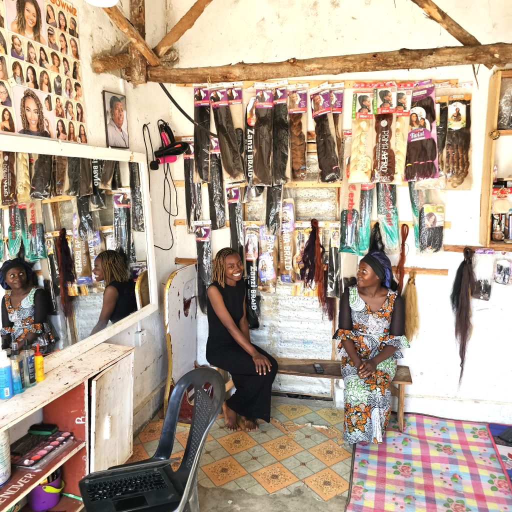 Two women sit in a hair salon next to a wall full of hair extensions.