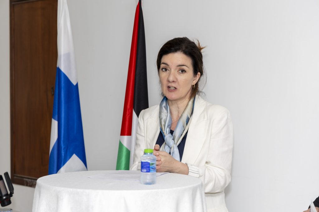 A woman stands at a table and speaks. Flags of Finland and Palestine in the background.