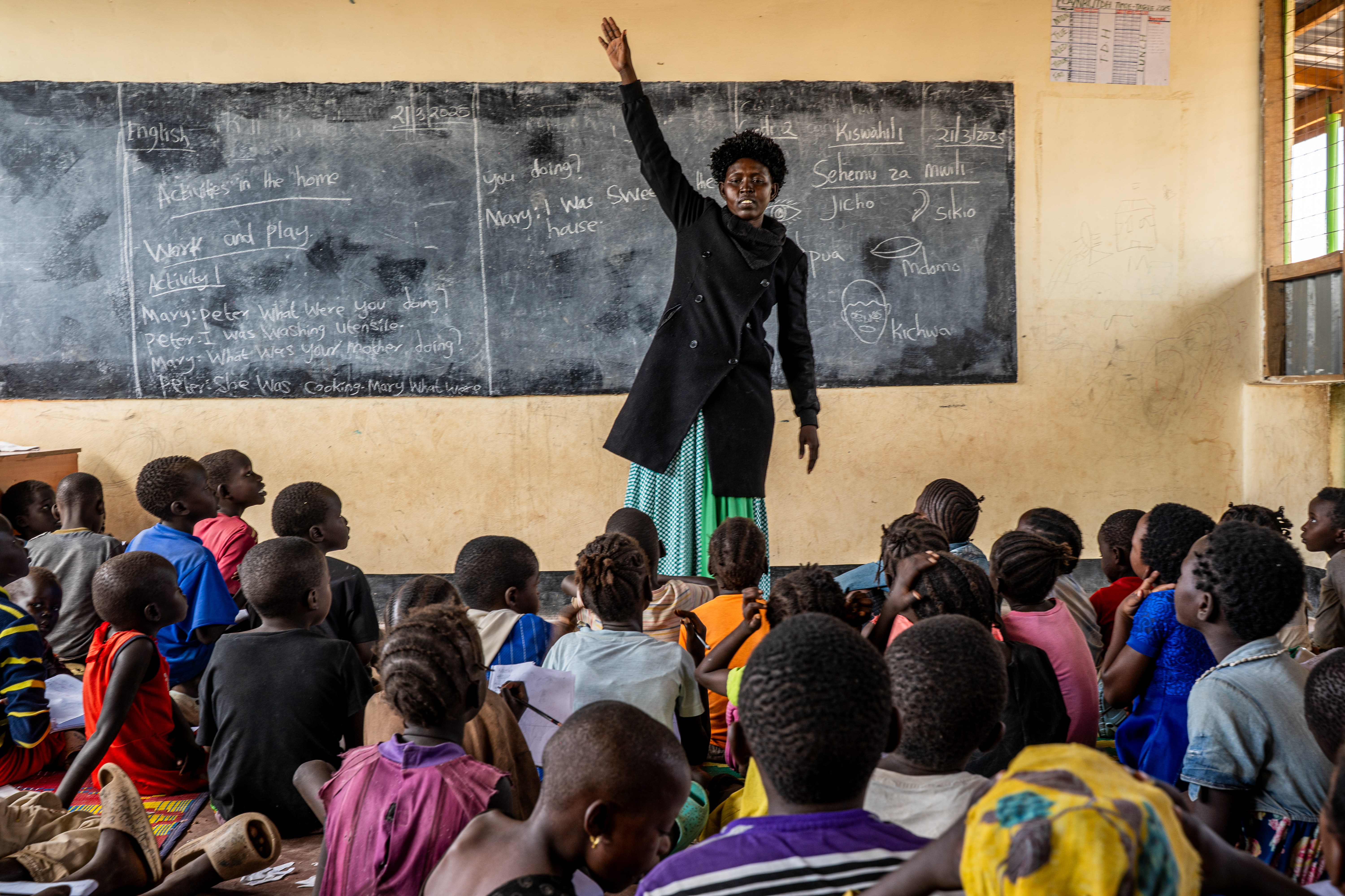 A woman stands in front of a blackboard and raises her hand. Children in front of her sit on the floor.