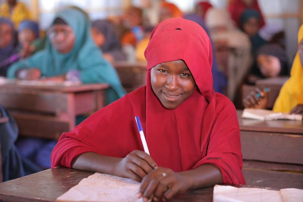 A smiling girl in a red hijab looks up from her desk in a classroom