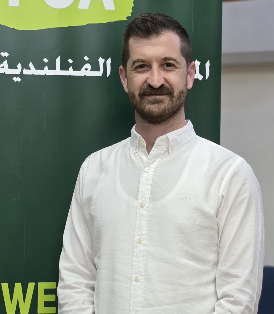 Person wearing a white shirt standing in front of a green Finn Church Aid banner with white Arabic text and yellow lettering, with a neutral indoor background including a brown door and blue seating.