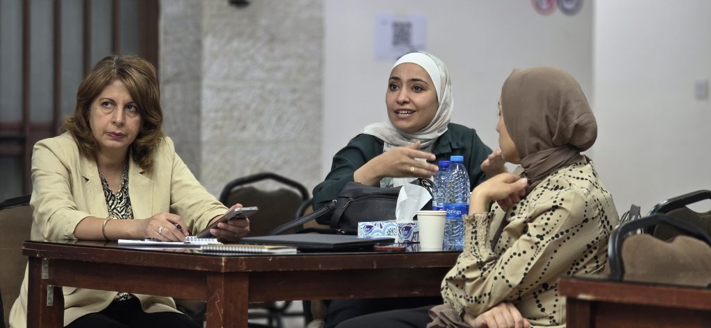 Three participants seated around a dark brown wooden table with notebooks, a laptop, a white cup, a tissue box with blue pattern, and water bottles, with one participant in a beige blazer holding a phone and others in green and beige clothing engaged in discussion, in a room with a beige wall and chairs.