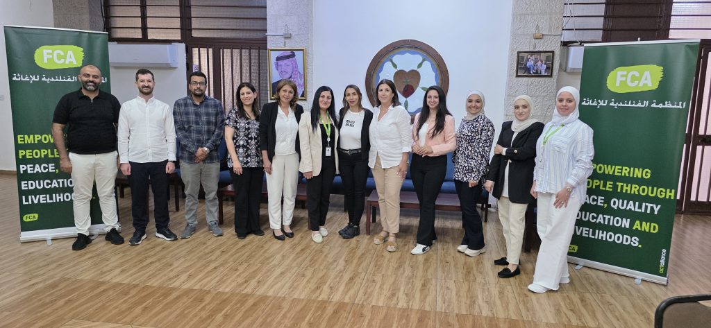 Group of participants standing on a wooden floor between two green Finn Church Aid banners with white Arabic text and yellow lettering, with a beige stone wall, framed pictures, and brown window panels in the background.