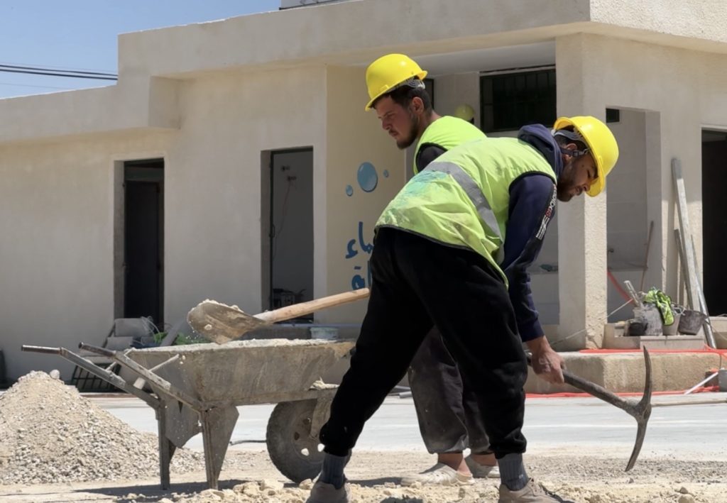Two local construction workers wearing yellow safety helmets and high-visibility light green safety vests carry out rehabilitation works at Attallah Saleh School in Al-Furqlus, Homs Governorate. One worker is using a pickaxe, while a wheelbarrow, construction materials, and the renovated school building are visible in the background as part of efforts to improve accessibility and create a more inclusive learning environment for students.