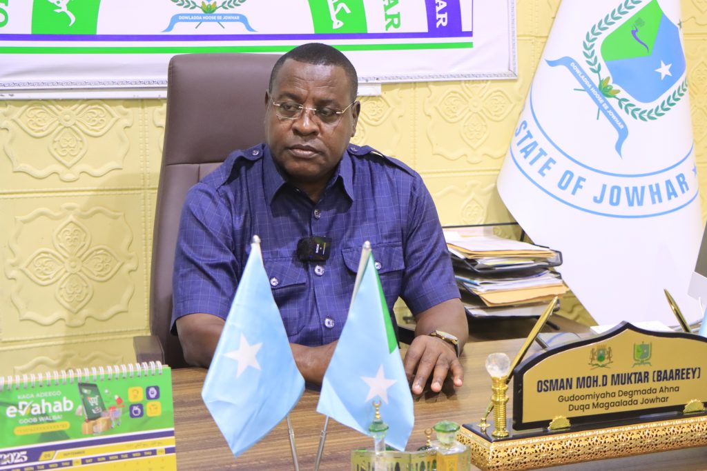 A man sits at an official looking desk. In front of him are two crossed flags of Somalia