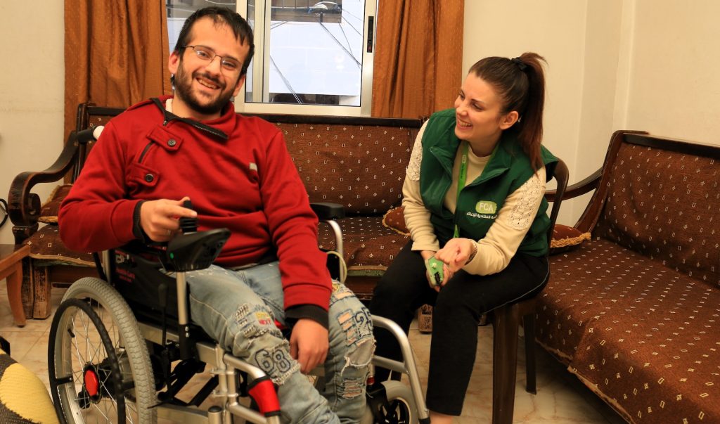 A smiling man in a wheelchair looks at the camera. To his left, sits an FCA staff member, sitting on a chair and looking towards him.