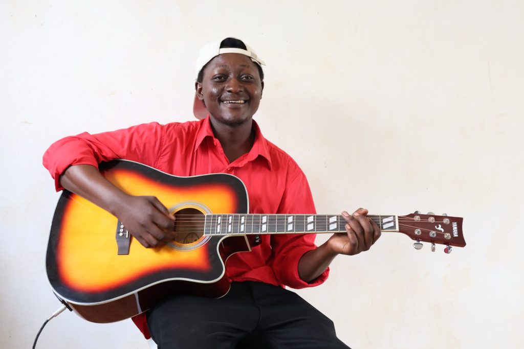 A smiling seated man in a baseball cap holds an acoustic guitar.