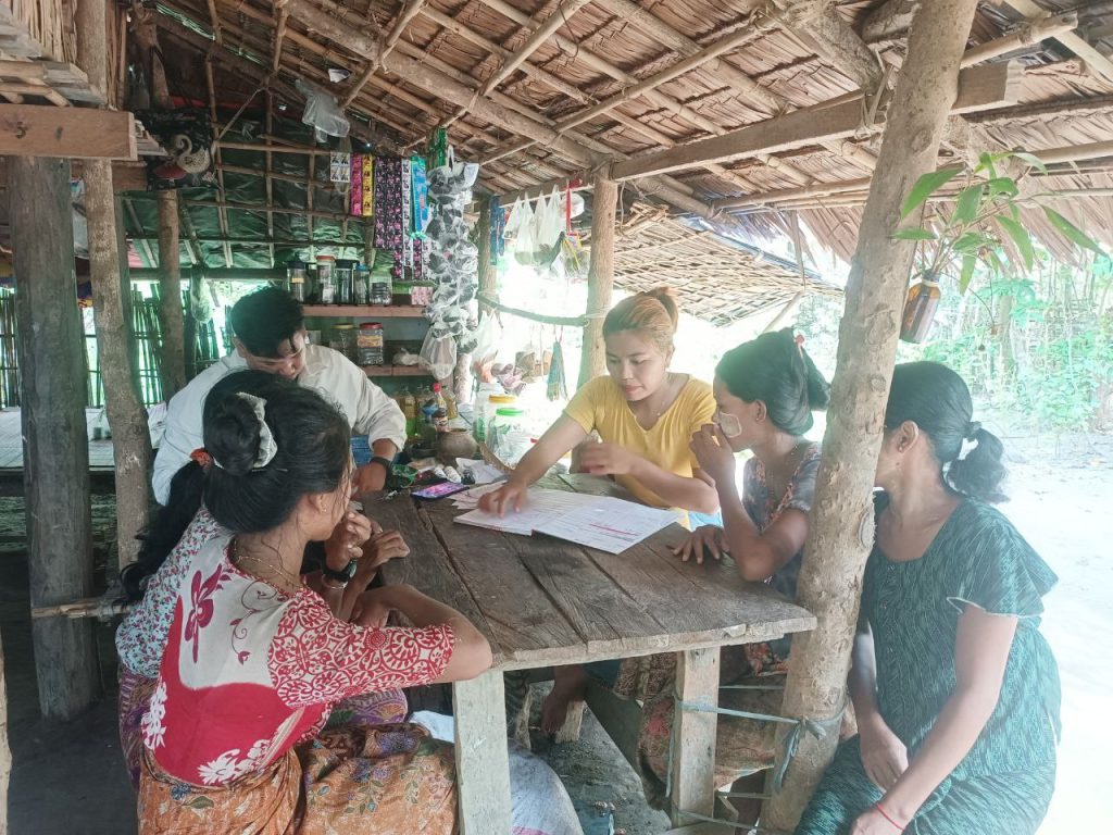 A group of women and a man sit on high stools and look at a book on a wooden table under a bamboo hut roof.