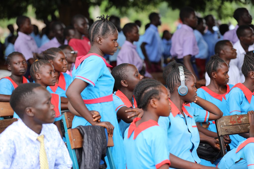 A girl in a blue and red school uniform stands in the midst of other similarly clad girls, who are seated outside and watching something offscreen