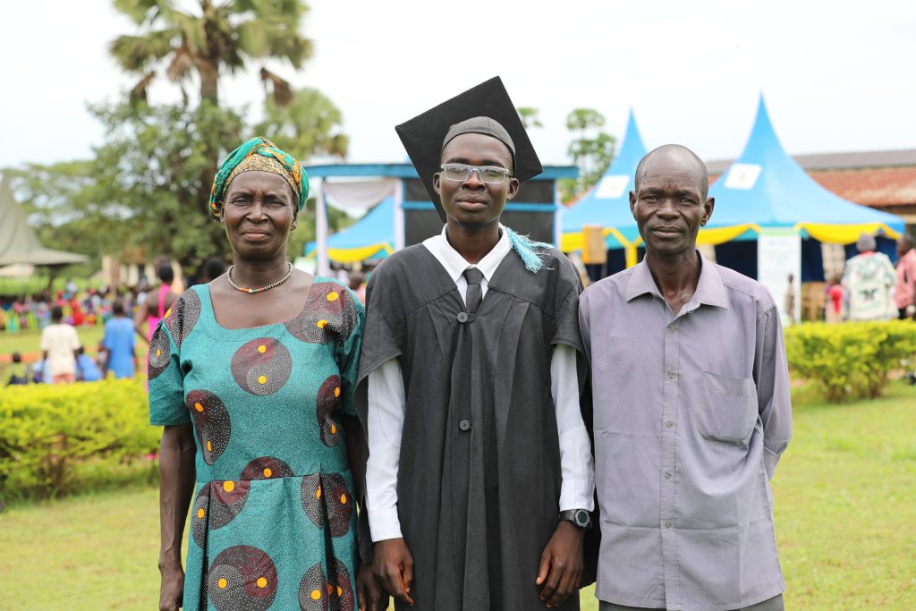 A young man in a graduate gown and cap stands outside with an older woman and man either side of him