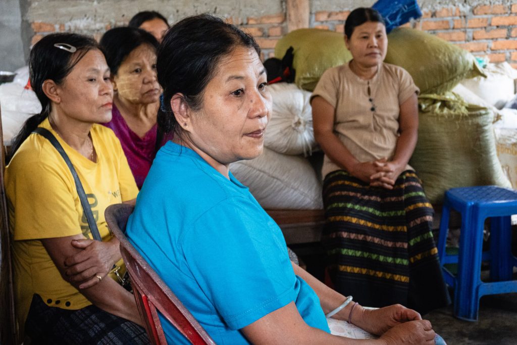 A woman in a blue shirt sits on a plastic chair in a warehouse. Other women sit behind her.