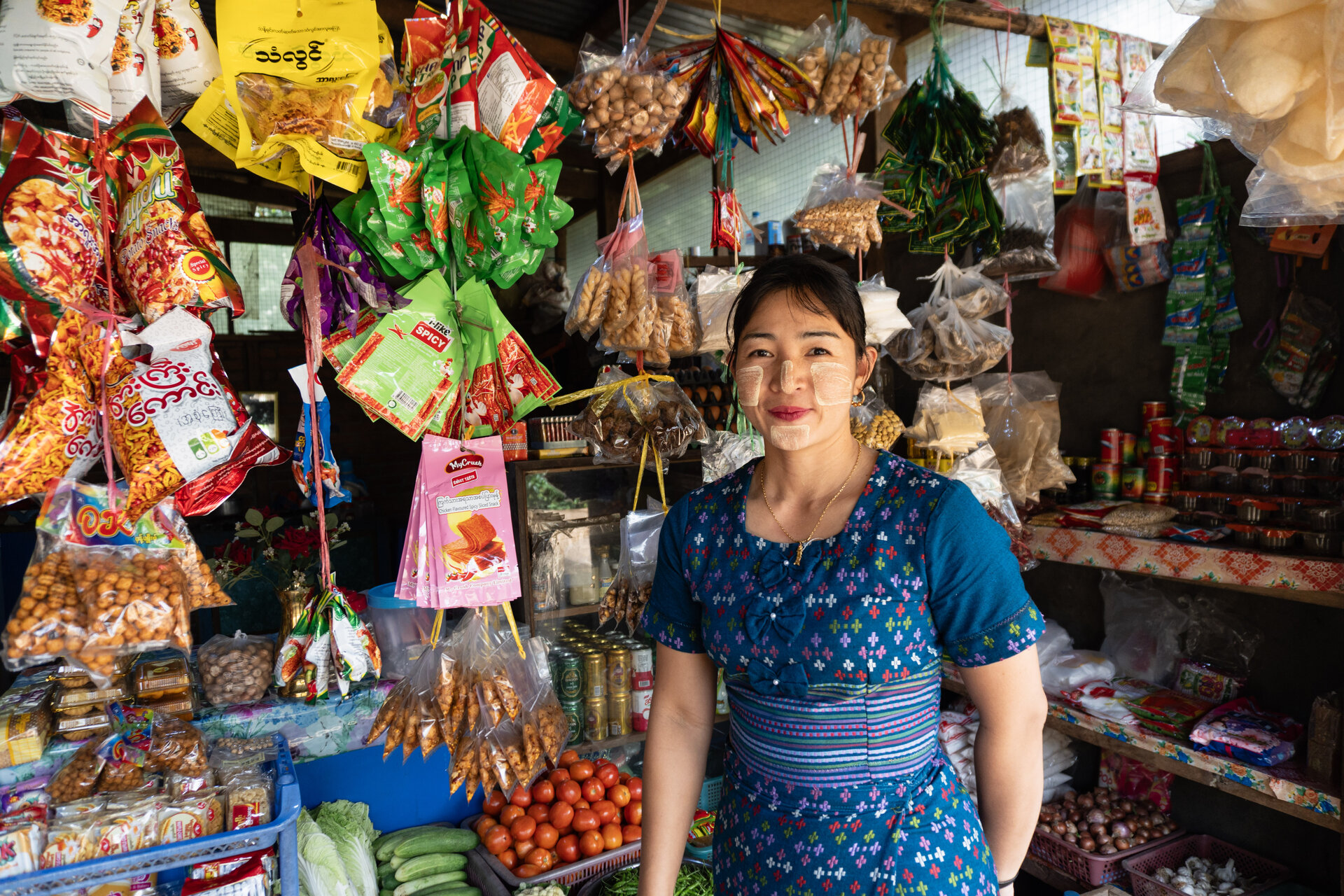 A smiling woman in a blue patterned dress and wearing traditional sunblock on her face stands in her shop. She is surrounded with different sorts of colourful groceries.