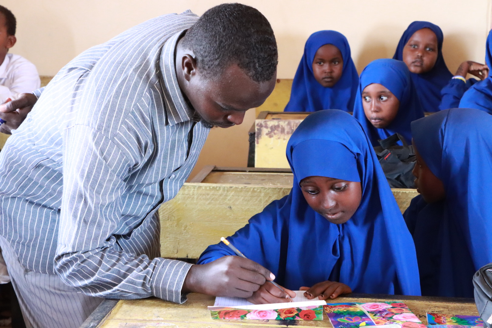 A male teacher leans over a schoolgirl in a blue hijab at a desk and helps her write into an exercise book