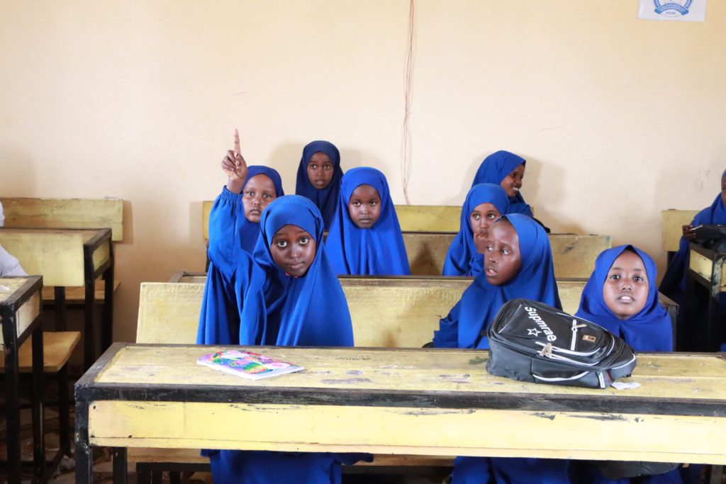 A schoolgirl in a blue hijab raises her hand sitting in a classroom with other similarly clad schoolgirls.