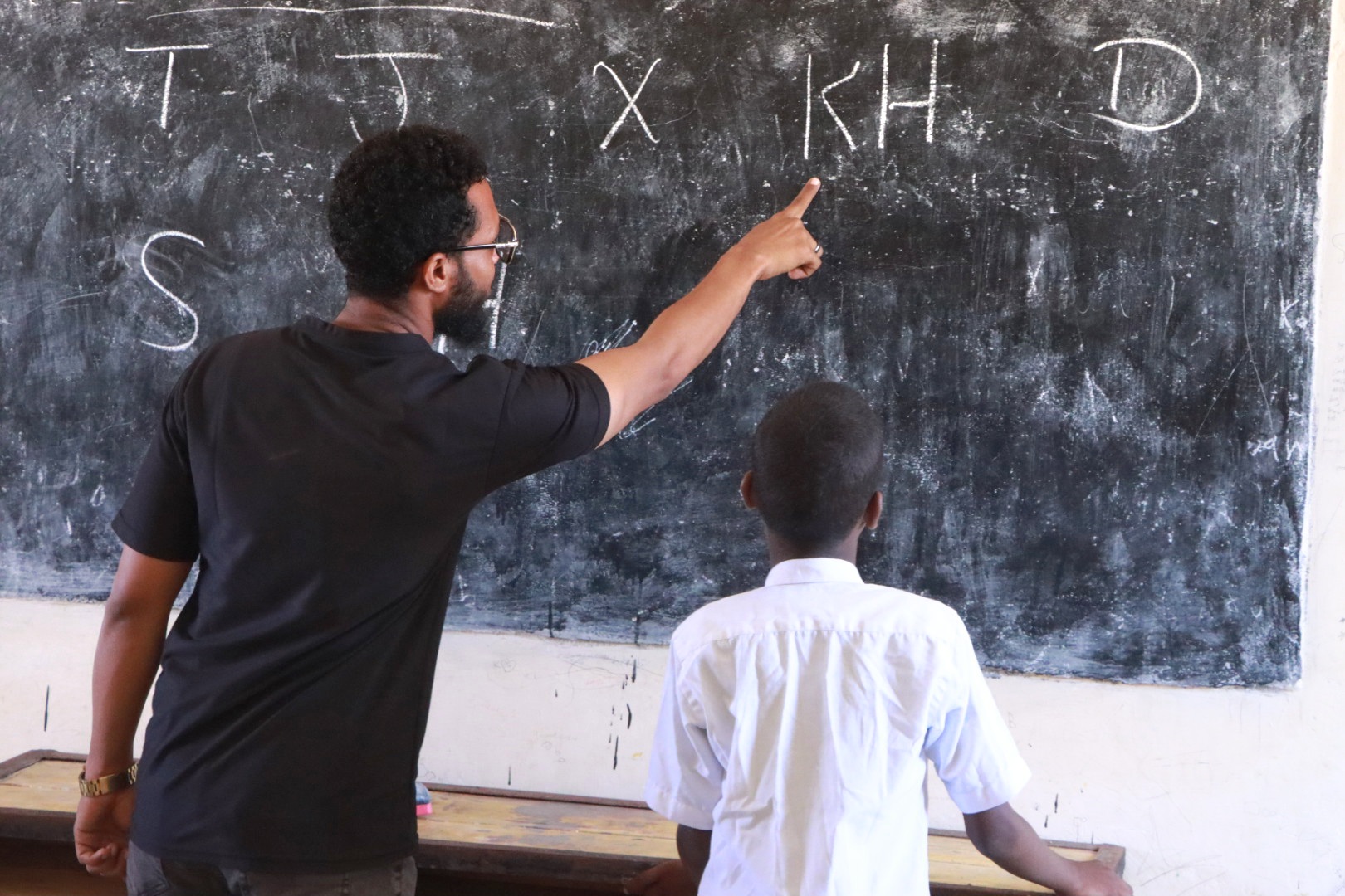A male teacher points to a Somali letter on a blackboard while a schoolboy in a white shirt looks on.